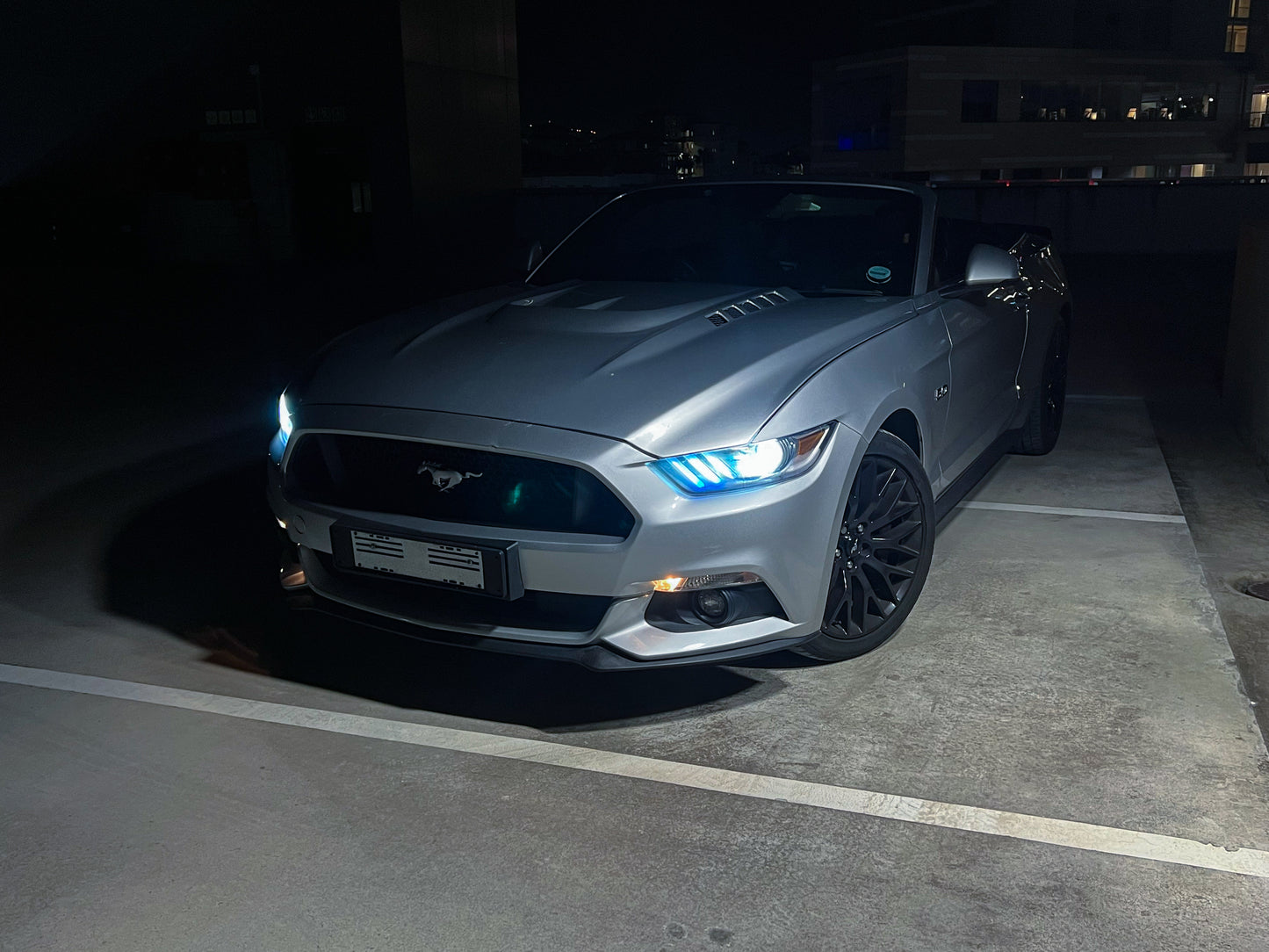 Gray Mustang car parked in a dark parking lot with illuminated headlights.