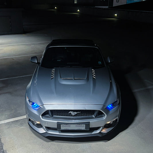 Gray Ford Mustang parked in a dark parking lot with illuminated headlights.